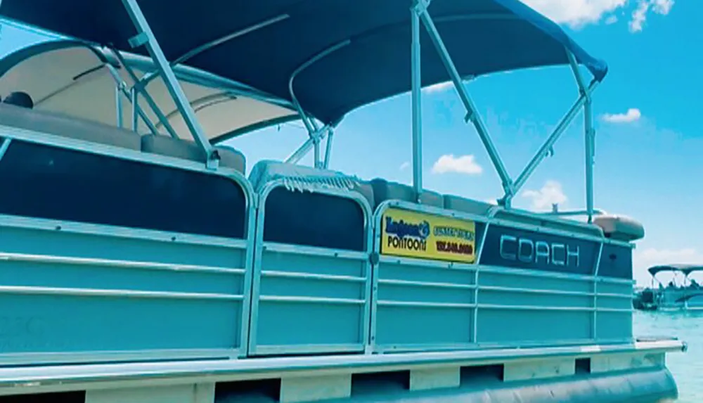 The image shows a side view of a pontoon boat equipped with a canopy moored on a body of water under a blue sky