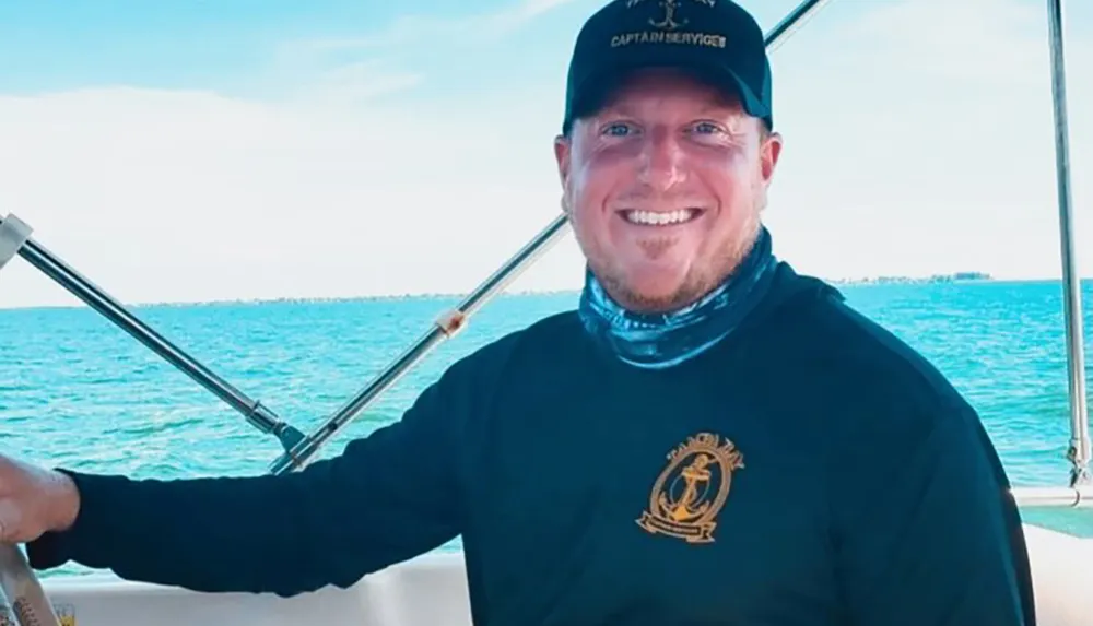 A smiling man wearing a cap and a long-sleeve shirt is steering a boat with clear blue skies and water in the background