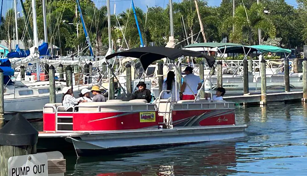 A group of people is enjoying a sunny day on a red pontoon boat at a marina filled with various boats and yachts