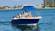 A group of people enjoy a sunny day on a blue motorboat on calm waters with coastal buildings in the background.