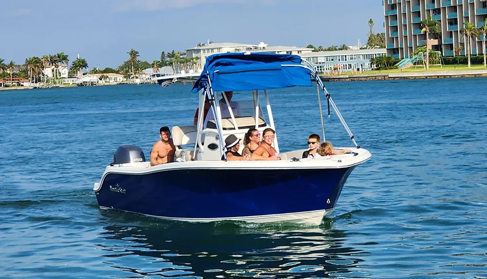 A group of people enjoy a sunny day on a blue motorboat on calm waters with coastal buildings in the background