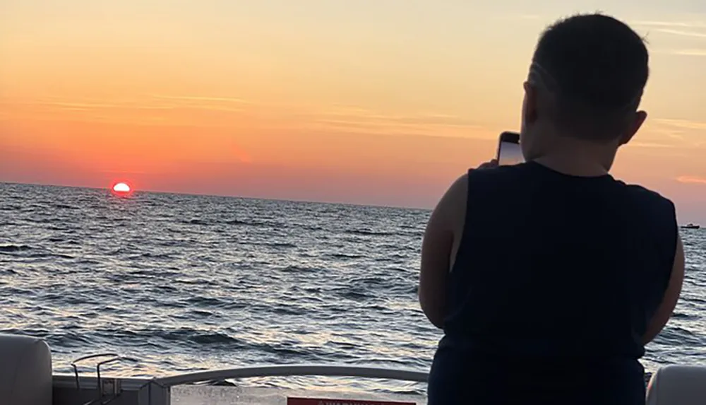 A person is capturing the sunset over the ocean with their smartphone from the deck of a boat