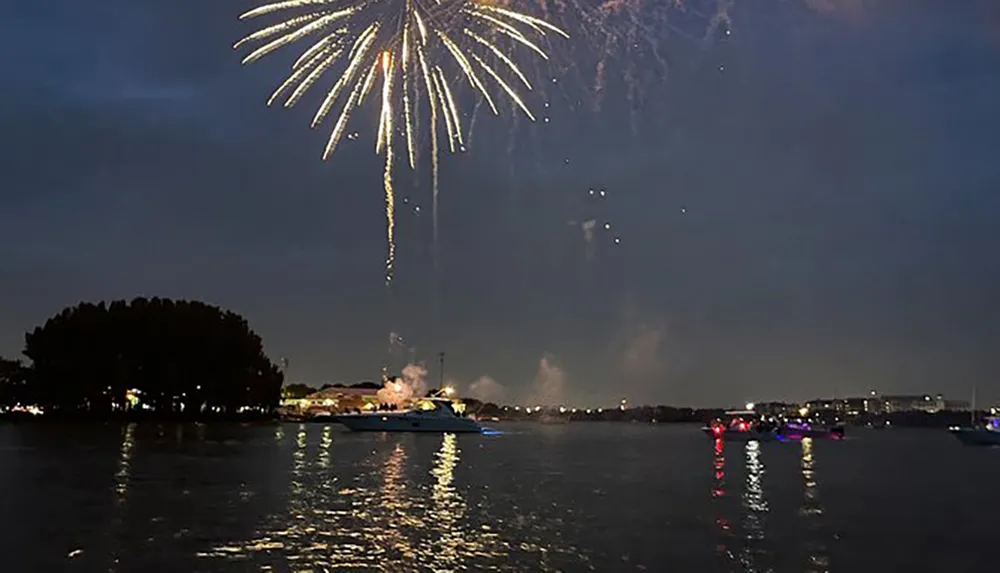 A firework bursts in the night sky above a body of water with boats reflecting lights on the rippling surface