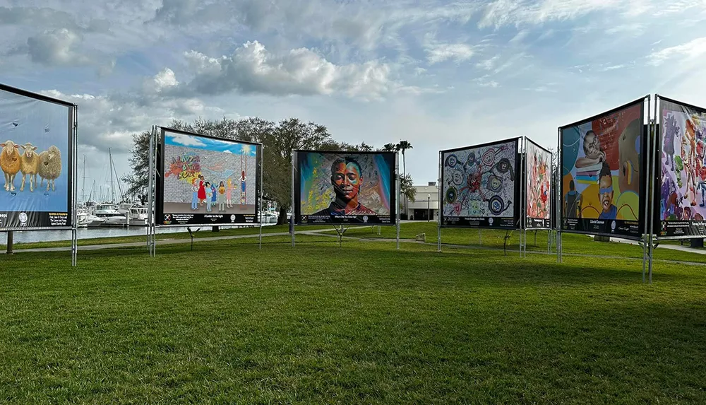 An outdoor exhibition featuring a series of large colorful artworks on display panels in a grassy area with a backdrop of trees and a marina