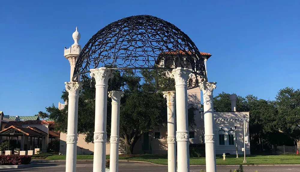 The image shows an ornate white gazebo with Corinthian columns supporting a decorative iron dome set in a sunny outdoor location