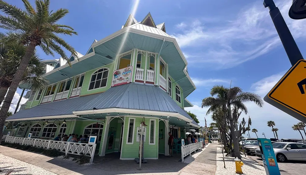 A charming two-story teal house with white trim and a wraparound porch nestled among palm trees under a blue sky appears to be a popular spot with patrons enjoying the outdoor seating area