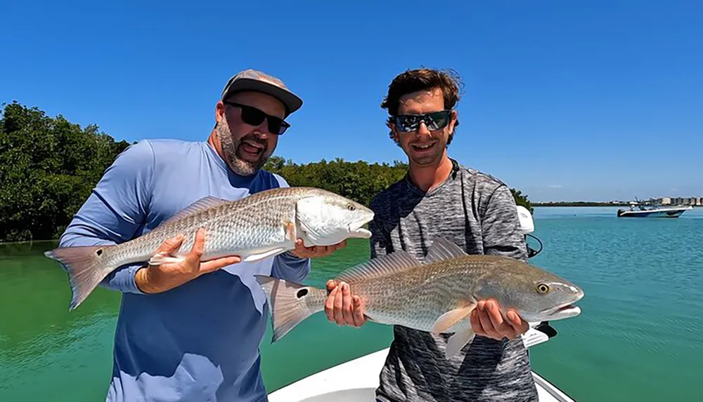 Two men are proudly displaying their catch of two large fish while standing on a boat with a clear blue sky and tranquil blue waters in the background