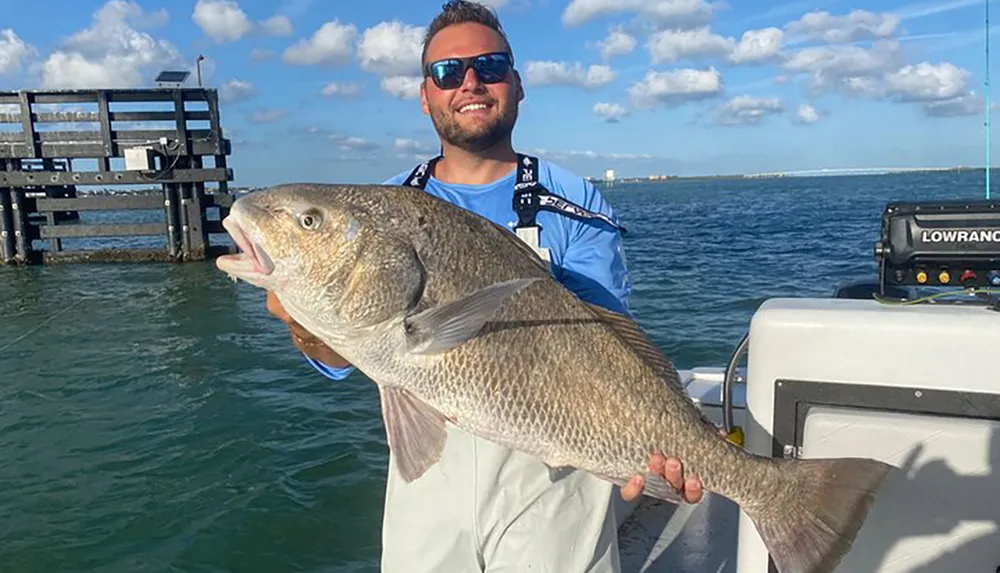 A smiling man is proudly holding a large fish on a boat with the ocean and a structure in the background