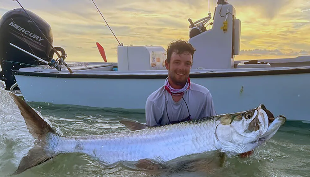 A smiling person is holding a large fish near a boat on the water during sunset