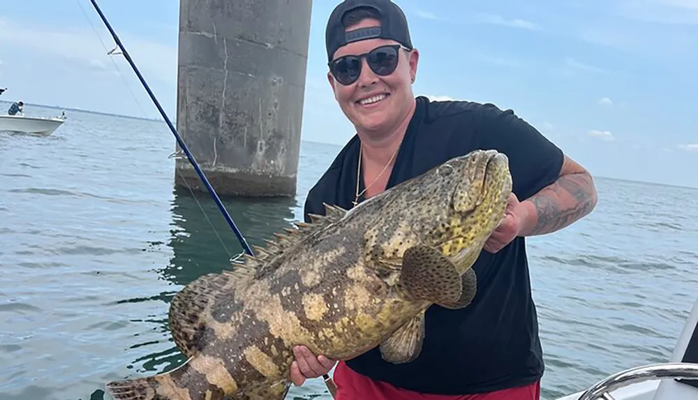 A person is proudly holding a large fish they caught while on a boat near a large concrete structure most likely a bridge pier with another boat visible in the background