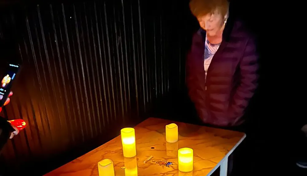 The image shows a person standing by a table with lit candles while another individual appears to be taking a photo with their smartphone in a dark setting