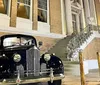 A vintage black car is parked in front of a building with grand steps and iron railings at night