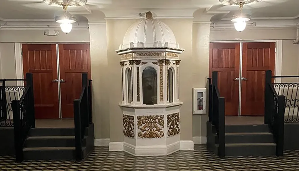 The image shows an ornate white elevator with decorative features flanked by two sets of wooden doors and black stair railings within an interior space featuring checkered flooring and a classic design aesthetic