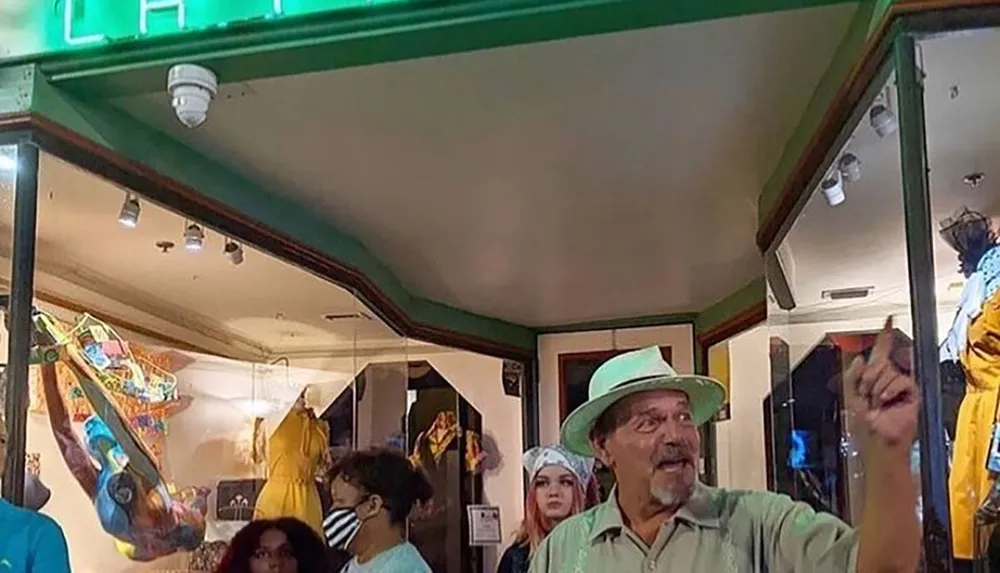 This image shows a man in a green hat gesturing with excitement inside a room with various people and colorful art or clothing in the background