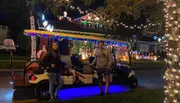 Two people are standing by a festively decorated golf cart in front of a house adorned with colorful Christmas lights at night.