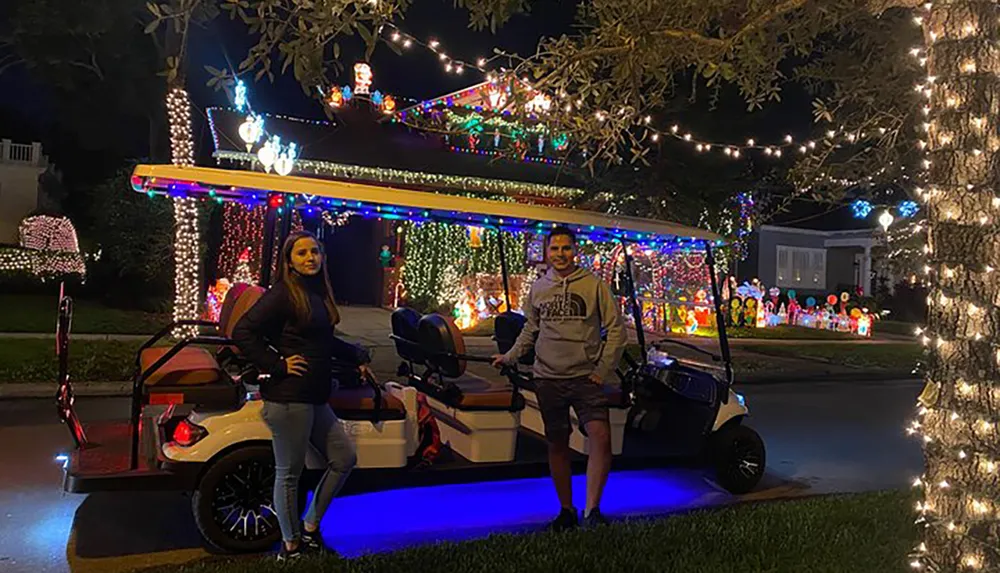 Two people are standing by a festively decorated golf cart in front of a house adorned with colorful Christmas lights at night