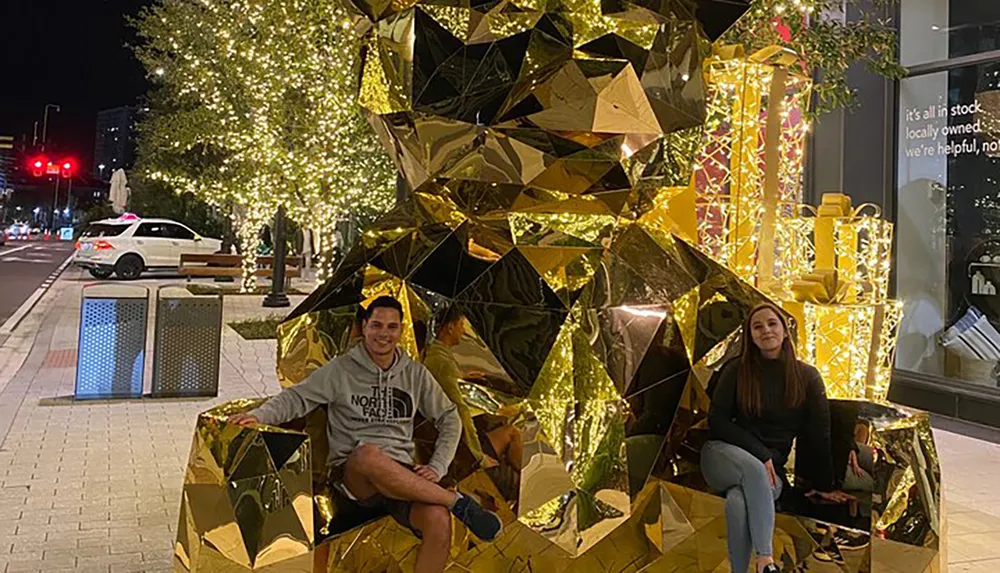 Two people are posing with smiles in front of a large reflective golden geometric sculpture surrounded by lights on a city street at night