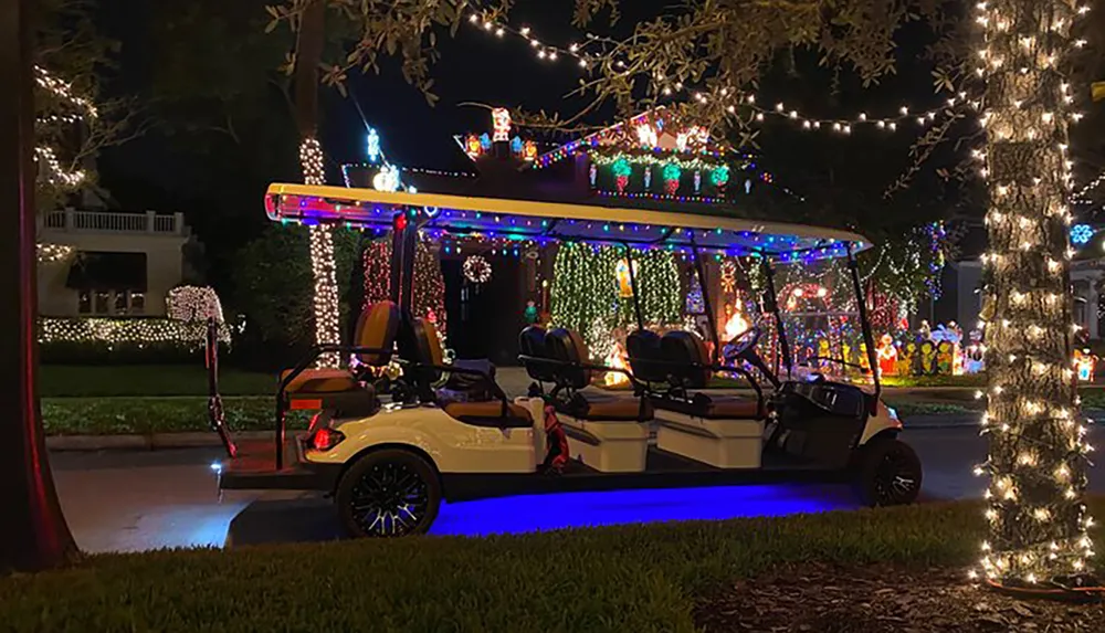 A golf cart adorned with festive lights is parked near a sidewalk lined with illuminated trees and a house extensively decorated with Christmas lights and decorations in the background at night