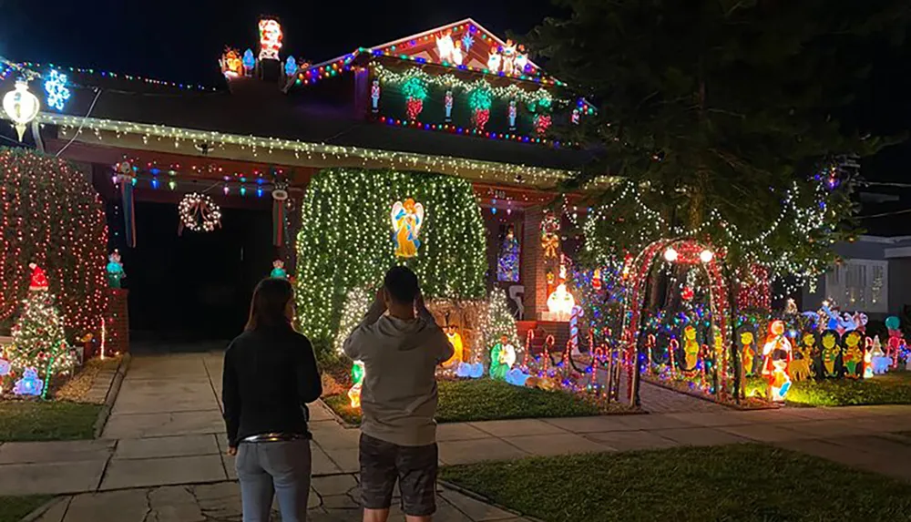 A decorated house with vibrant Christmas lights and figures is being admired by two people standing on the sidewalk at night