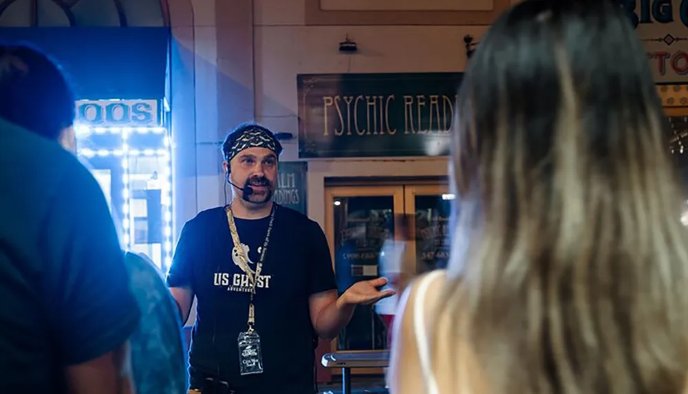 A man wearing a bandana and a microphone headset guides a tour in front of a psychic readings shop while gesturing to a person whose back is facing the camera