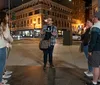 A group of people appear to be engaged in a conversation on a city street at night with illuminated buildings in the background