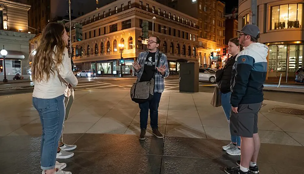 A group of people appear to be engaged in a conversation on a city street at night with illuminated buildings in the background