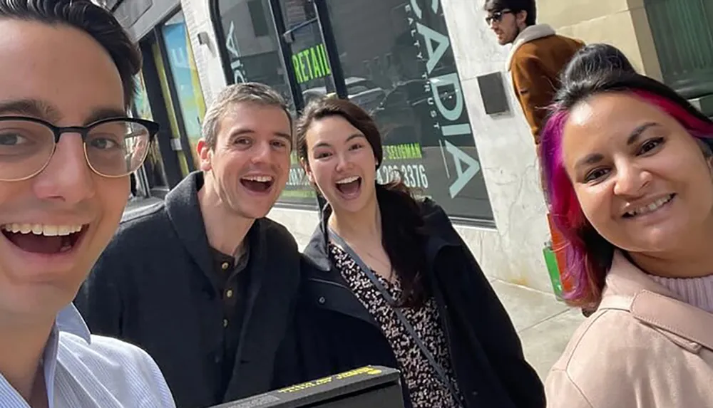 Four individuals are posing for a selfie on a sunny day with bright smiles and a city environment in the background