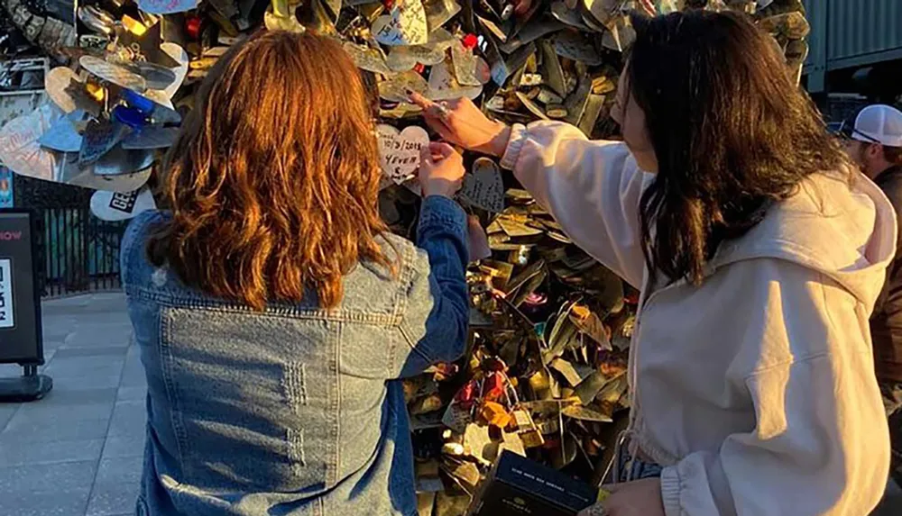 Two people are attaching a lock to a collection of locks on a metal structure a gesture often associated with making a wish or symbolizing love and commitment