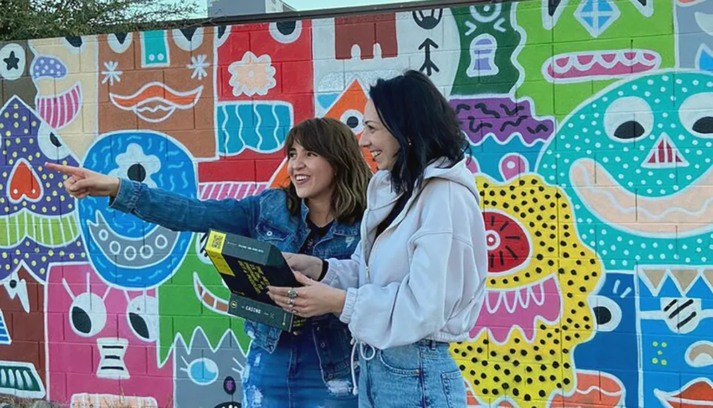 Two people are smiling and interacting with each other in front of a colorful mural-covered wall