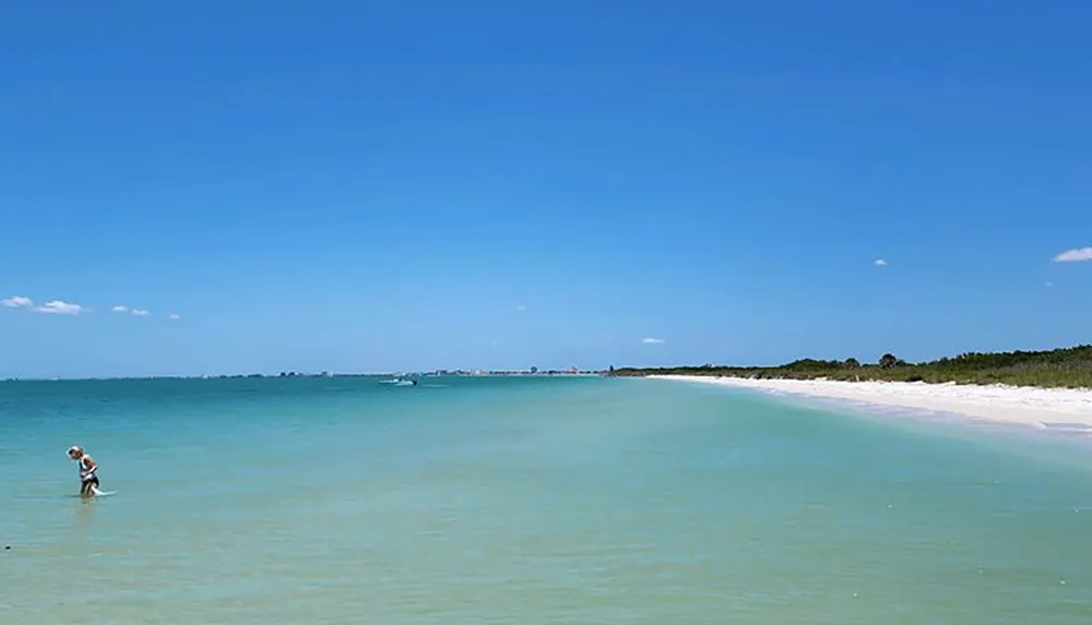 The image shows a person standing in shallow clear waters near the shore of a beautiful beach on a sunny day