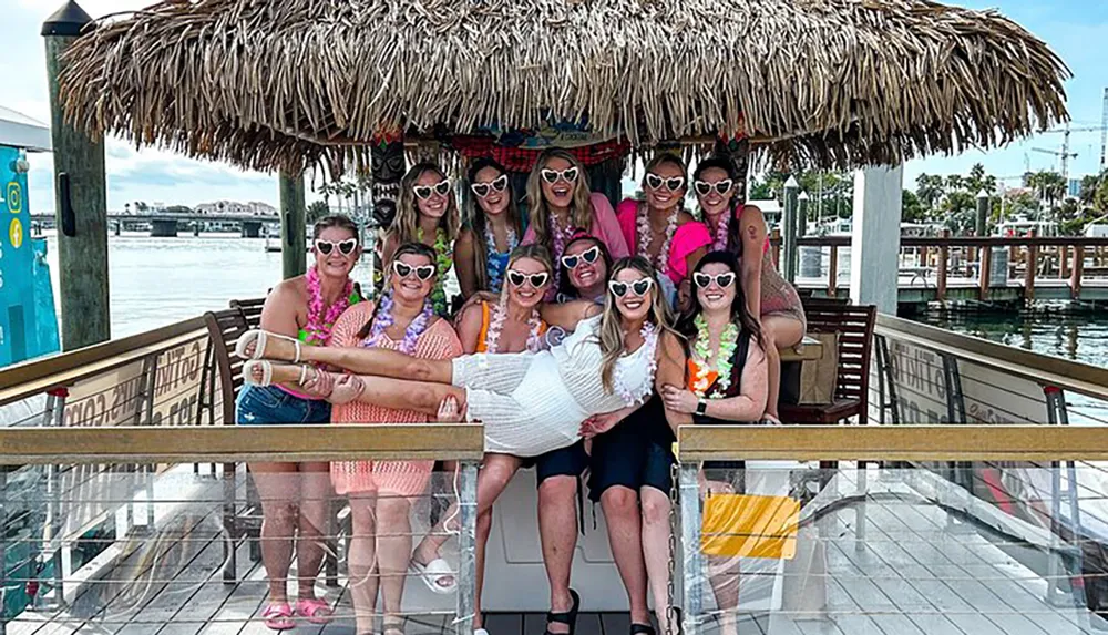 A group of smiling people likely friends or colleagues are posing for a photo together on a sunny day under a tiki hut-style structure with several of them wearing sunglasses and colorful leis