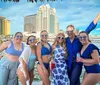 A group of smiling people wearing matching heart-shaped sunglasses is posing for a photo in a festive atmosphere by the waterfront with a city skyline in the background