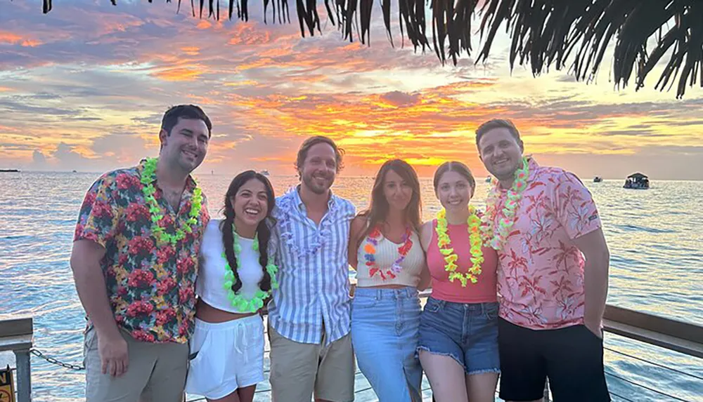 A group of six people wearing tropical-themed clothing and leis are posing for a photo with a vibrant sunset over the ocean in the background