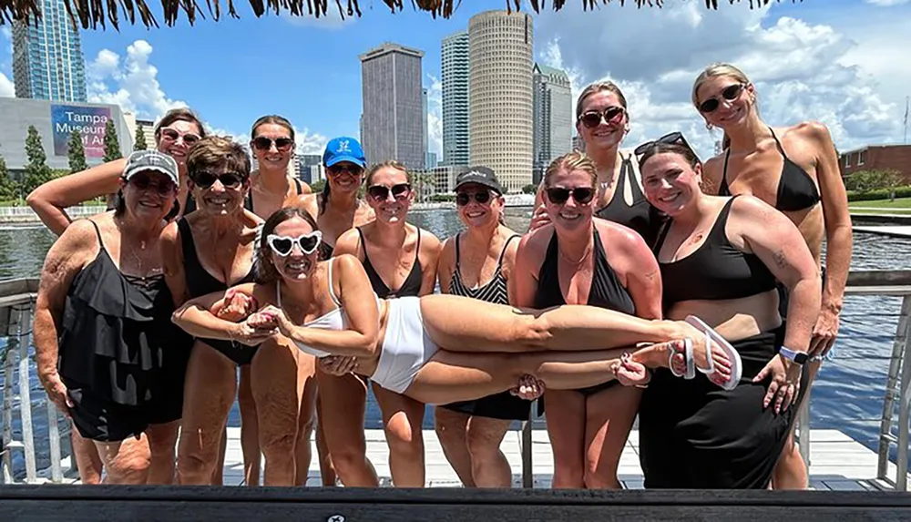 A group of cheerful people stand by the waterfront on a sunny day posing for a fun photo with one person playfully being lifted in front