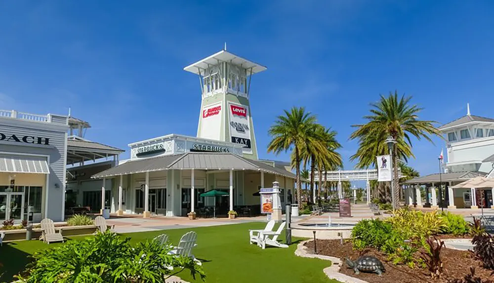 The image shows an outdoor shopping center with palm trees grassy areas and a variety of store fronts under clear blue skies