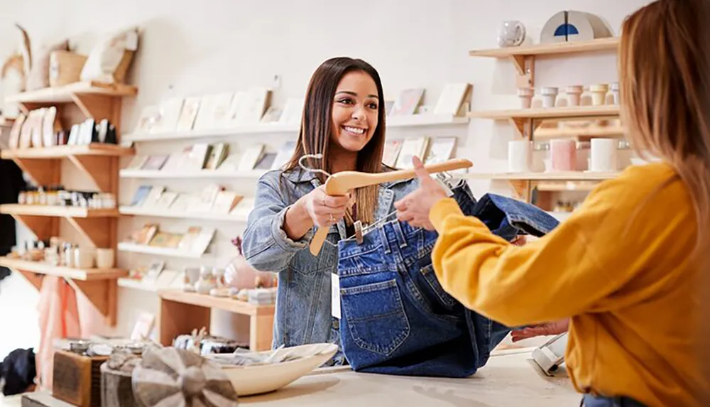 A smiling salesperson is handing a clothing item on a hanger to a customer in a boutique store