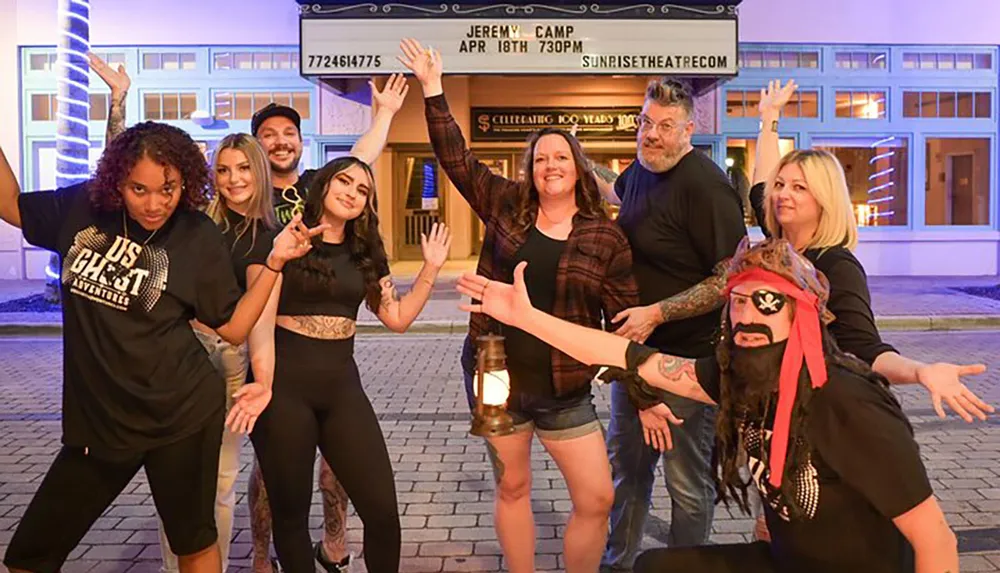 A diverse group of people are posing joyfully in front of a theater marquee with one person dressed up as a pirate kneeling in front