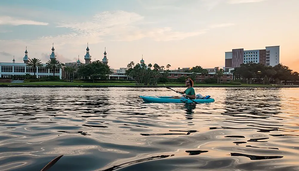 A person is kayaking on calm waters with a scenic backdrop of a building with multiple spires during sunset