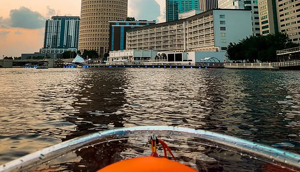 The image shows a city waterfront viewed from a boat with modern buildings lining the shore against an evening sky