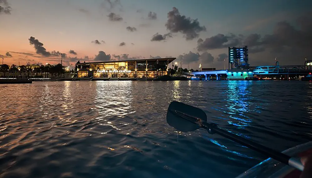 The image captures a serene evening view from the perspective of a person kayaking on the water near an illuminated building and bridge under a dusk sky