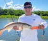 A smiling person is holding a caught fish on a sunny day with a fishing rod and reel in the background indicating a successful fishing trip