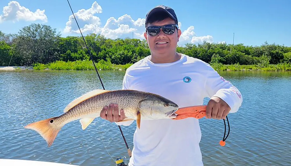 A smiling person is holding a caught fish on a sunny day with a fishing rod and reel in the background indicating a successful fishing trip