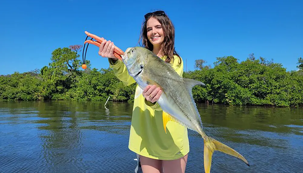 A woman is holding a large fish with a smile standing in front of a body of water with lush greenery in the background