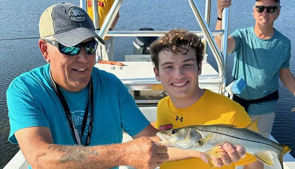 Two smiling people on a boat are proudly displaying a fish theyve caught with another individual cheerfully observing in the background