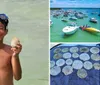 A joyful child wearing a snorkel mask holds up a sand dollar and a seashell while standing in shallow water at the beach