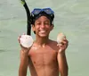 A joyful child wearing a snorkel mask holds up a sand dollar and a seashell while standing in shallow water at the beach