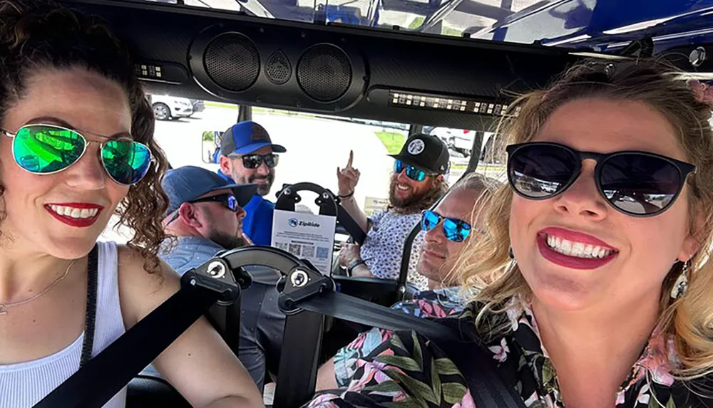 A group of smiling people in sunglasses takes a group selfie while riding in a golf cart