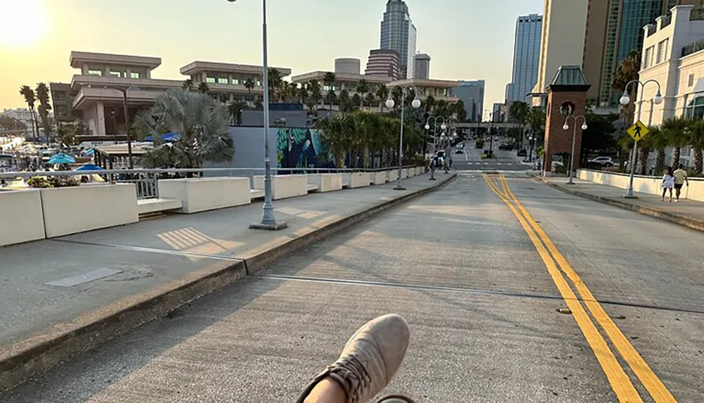 The image shows a persons perspective with their foot in the foreground looking down a sunny urban street lined with palm trees and lampposts leading towards high-rise buildings in the distance