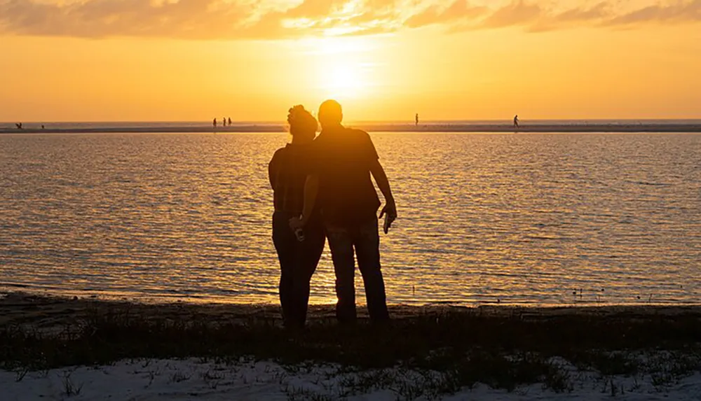 Two silhouetted figures stand closely together watching a stunning sunset over a tranquil ocean with other people visible in the distance