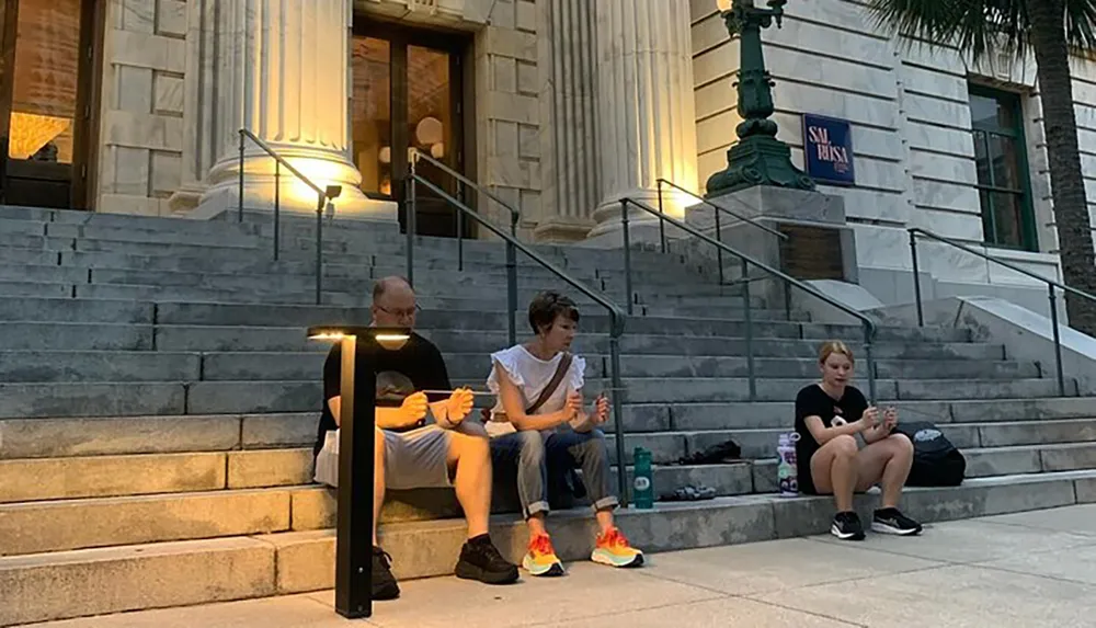 Three people are sitting on the steps of a building each engrossed in their own phone while the evening sets in